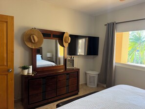 A bedroom with a wooden dresser, a mirror, a television, a window with curtains, and a bed with a patterned bedspread. - Luxury Cabo Condo with Ocean Views Overlooking Cabo Real Golf Course (San José del Cabo)