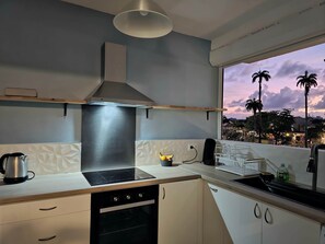 A modern kitchen with a built-in oven, a range hood, and a window overlooking a cityscape at dusk. - Amandine - vue mer Pointe-à-Pitre (Pointe-a-Pitre)