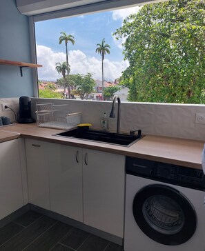A modern kitchen with a built-in dishwasher, a coffee maker, and a window overlooking a cityscape with palm trees. - Amandine - vue mer Pointe-à-Pitre (Pointe-a-Pitre)