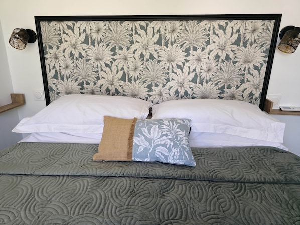 A bed with a patterned headboard, two pillows, a textured blanket, and wall-mounted lamps. - Amandine - vue mer Pointe-à-Pitre (Pointe-a-Pitre)