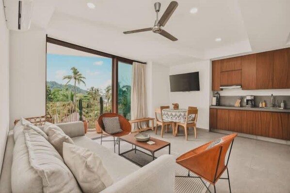 Een moderne woonkamer met een bank, salontafel en fauteuils, een eethoek met een tafel en stoelen, een keuken met houten kasten en een flatscreen televisie, en een balkon met uitzicht op palmbomen.