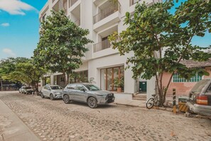 A multi-story building with balconies, parked cars, and trees along the street. - Balconies Tranquillo at La Esquina: San Pancho 2BR, Ocean Views, Near Beach (San Pancho)