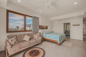 A bedroom with a sofa, a bed, a ceiling fan, and a large window with a view of palm trees. - Balconies Tranquillo at La Esquina: San Pancho 2BR, Ocean Views, Near Beach (San Pancho)