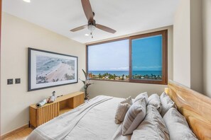 A bedroom with a bed, a ceiling fan, a window with a view of the beach, and a framed picture on the wall. - Vista Panoramic at Caivama: Sayulita 2BR, Plunge Pool & Ocean View (Sayulita)