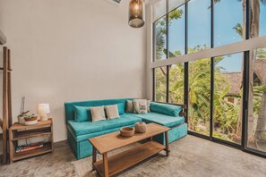 A room with a teal sofa, a wooden coffee table, a side table with a lamp, and a large window offering a view of greenery. - Campanilla at Ipomea: Sayulita Loft w/ Terrace, Lap Pool, Jungle (Sayulita)