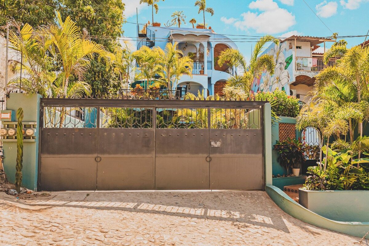 A gated entrance with a cobblestone path leading to a building with balconies and tropical plants.