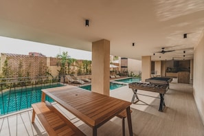 Uma área de piscina ao ar livre com mesa de madeira e mesa de futebol, cercada por espreguiçadeiras e vista para a piscina.