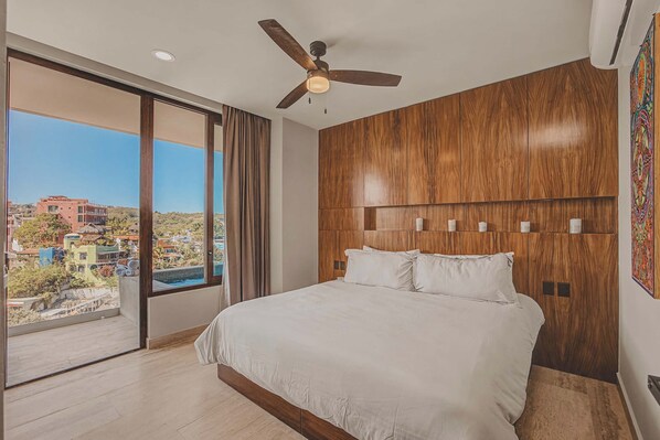A bedroom with a large bed, a ceiling fan, and a view of buildings through a large window. - El Umbral at Caivama: Sayulita 2BR, Yoga Terrace & Plunge Pool (Sayulita)