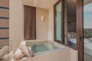 Ein Balkon mit einem gläsernen Whirlpool, Holzklappläden und einem Bett, das durch eine Glastür sichtbar ist.