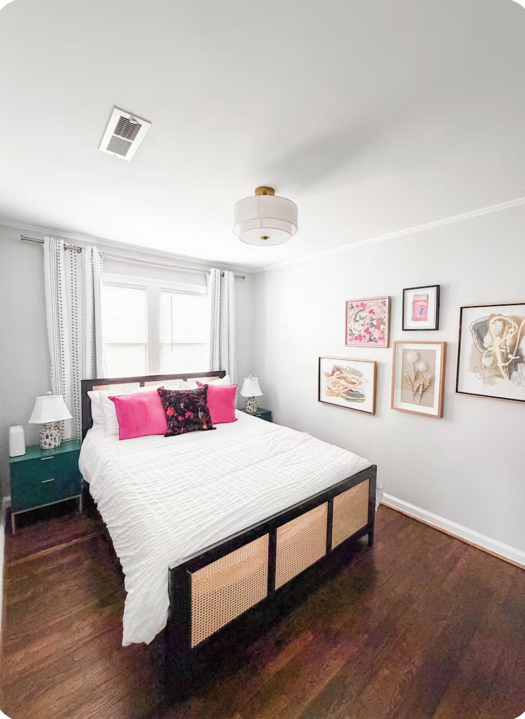 Une chambre à coucher comprenant un lit, une table de chevet, une lampe, une fenêtre avec des rideaux, une bouche d’aération au plafond et des œuvres d’art murales.