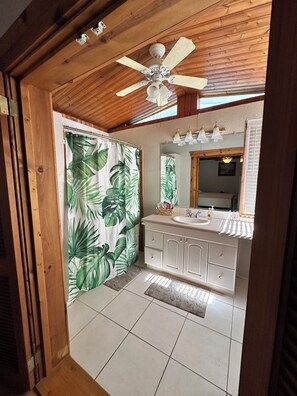 Une salle de bain avec un plafond en bois, un ventilateur de plafond, un rideau de douche orné de motifs de feuilles, un lavabo blanc et un miroir.