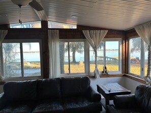 Ein Zimmer mit Ledersofa, einem kleinen Tisch und großen Fenstern mit Blick auf einen See und eine Anlegestelle.