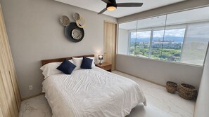 A bedroom with a bed, a nightstand, a wall-mounted light, a ceiling fan, a large window with a view, and two wicker baskets. - New Listing (Bucerías)