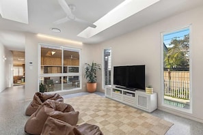 A spacious living area with a large window, a flat-screen TV, and a ceiling fan. - Paradise Club by Coast Hosting (Wamberal)