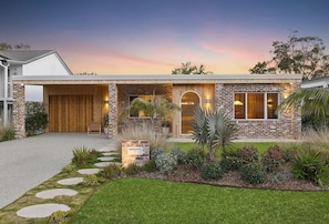 A modern two-story house with a brick facade, a wooden garage door, and a well-maintained front yard with stepping stones and various plants. - Paradise Club by Coast Hosting (Wamberal)