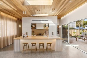 A modern kitchen with a central island, bar stools, and a view to the outside. - Paradise Club by Coast Hosting (Wamberal)