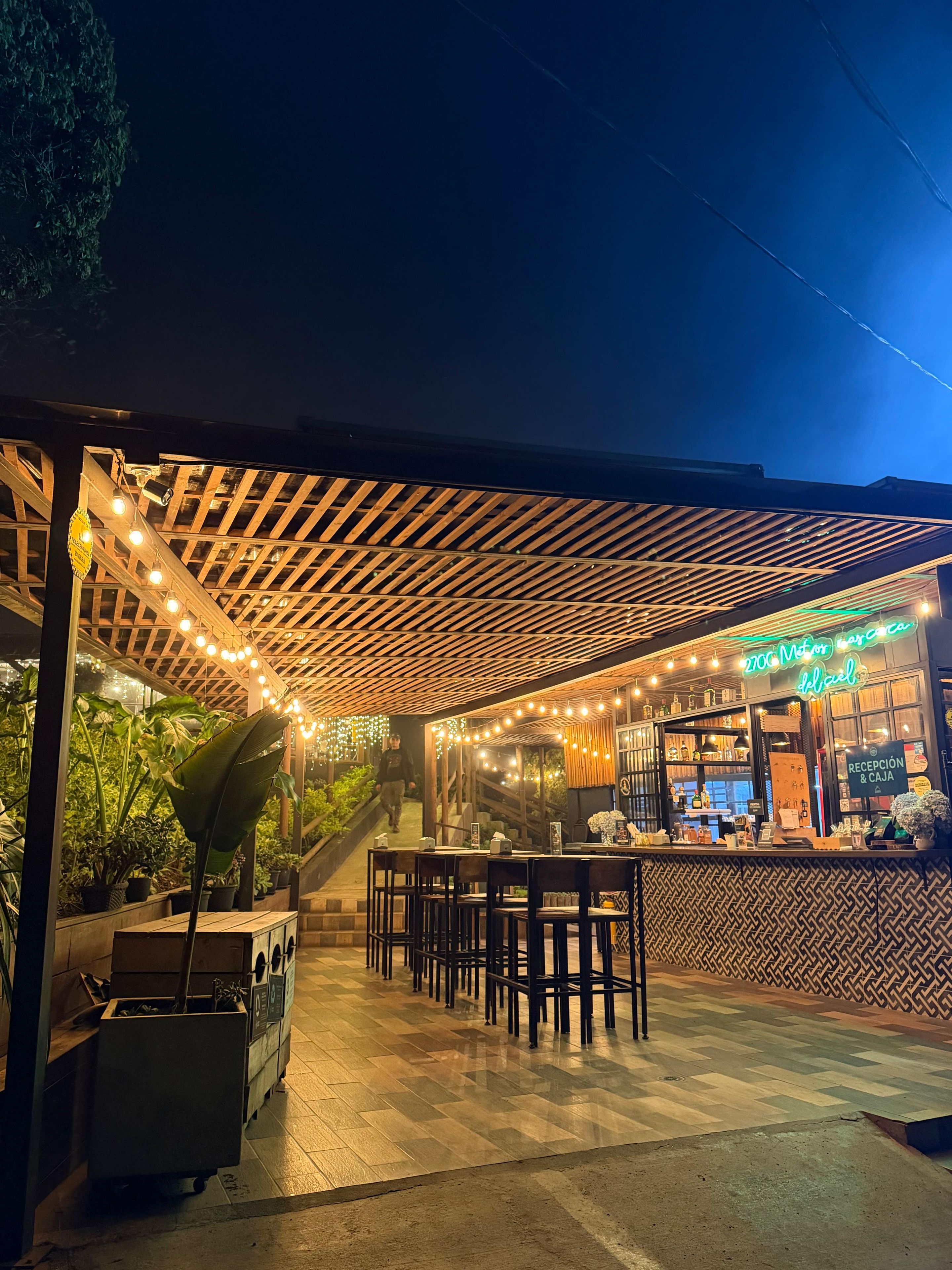 Un área de estar al aire libre con mesas y sillas, una pérgola de madera, luces colgantes y una zona de bar con un letrero de neón.