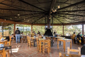 Un café en plein air avec des tables et des chaises en bois, une grande cheminée noire et une structure de toit en métal.