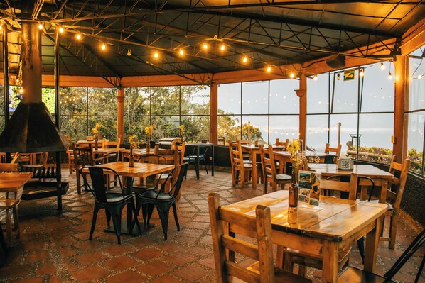 An outdoor dining area with tables and chairs, a large window overlooking greenery, and string lights on the ceiling.