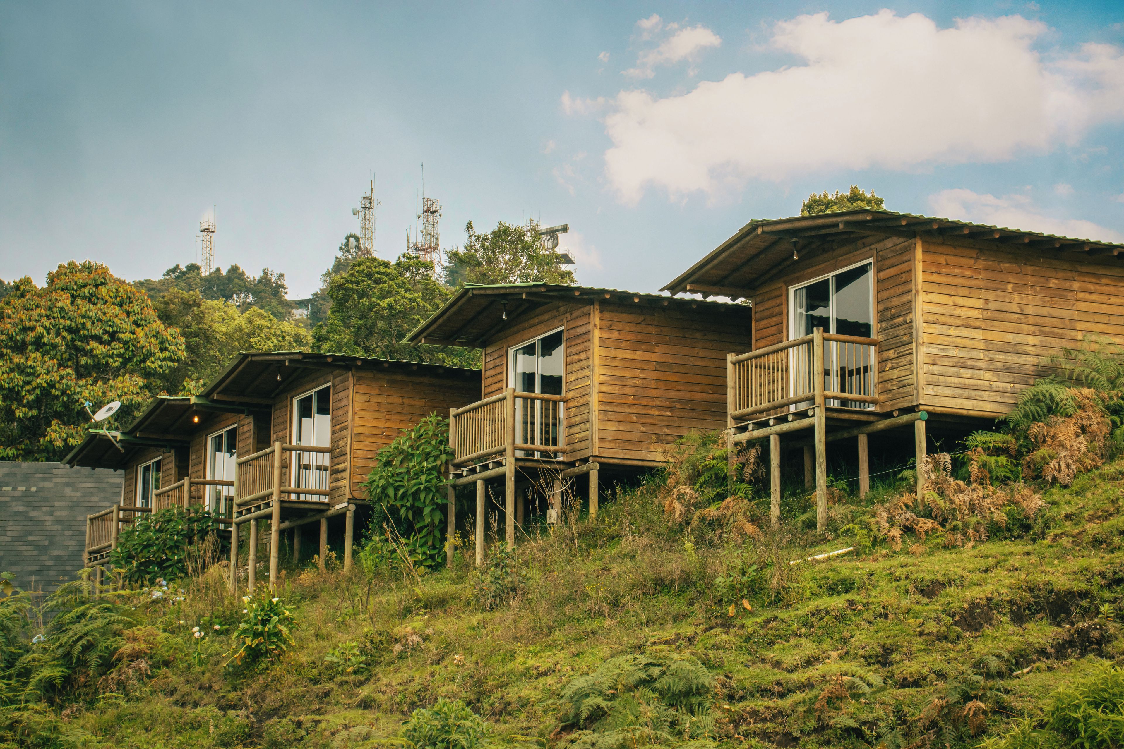Una hilera de cabañas de madera con balcones, ubicadas en una ladera con abundante vegetación y un cielo despejado.