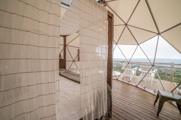 Una habitación con vista a un paisaje, piso de madera y cortina blanca.
