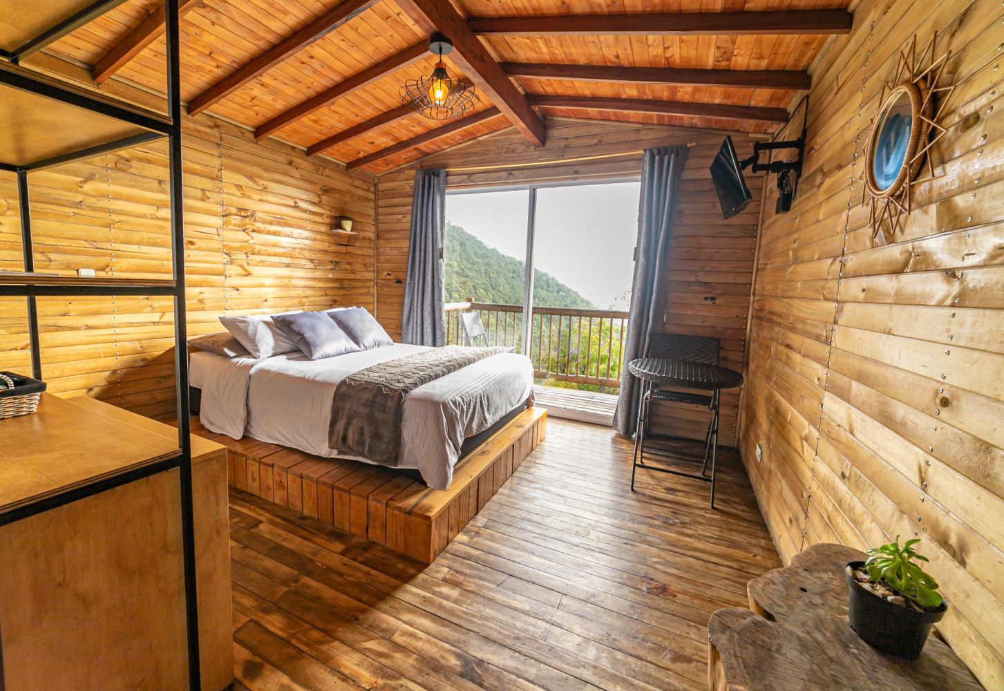 A wooden cabin room with a bed, a small table, and a balcony with a view of the mountains.