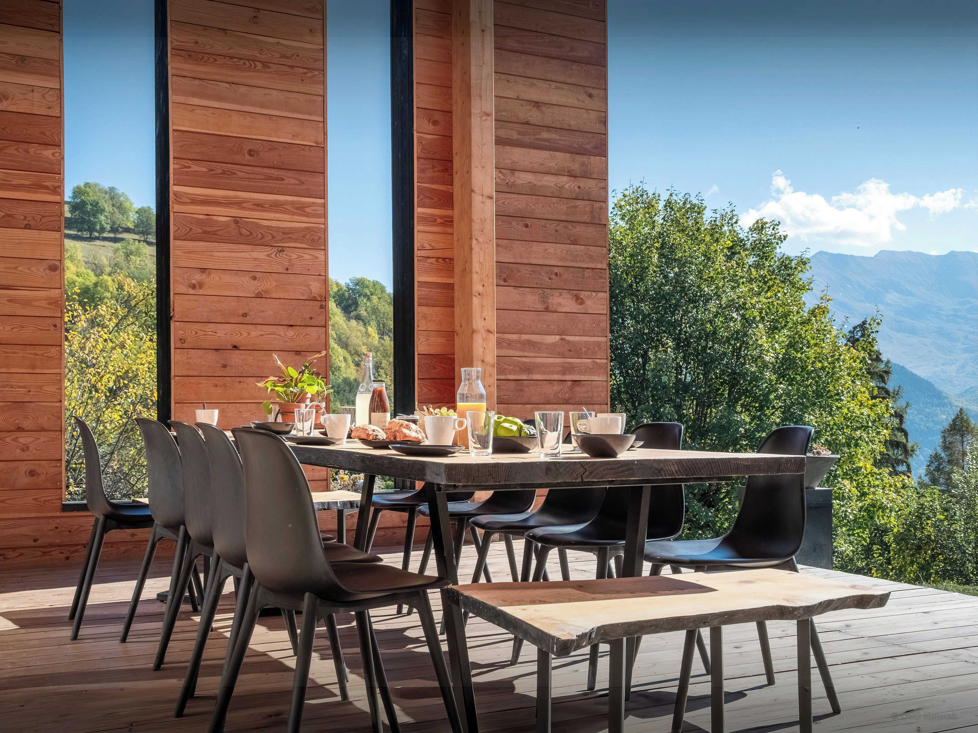 Un espace de restauration en plein air avec une table et des chaises en bois, une bouteille de vin et une vue sur un paysage montagneux.