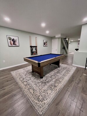 Una mesa de billar en una habitación con alfombra estampada, un atril para billar y dos cuadros en la pared.