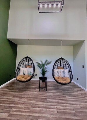 Zwei Hängesessel mit Kissen, ein kleiner Tisch und ein Blumentopf in einem Raum mit Holzboden und einer grünen Akzentwand.