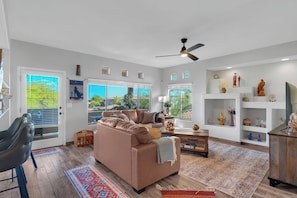A modern living room with a brown leather sofa, a ceiling fan, and a view of a pool through large windows. - Oro Valley 2BR Condo with Pool-View Balcony, Shared Pool & Hot Tub (Oro Valley)