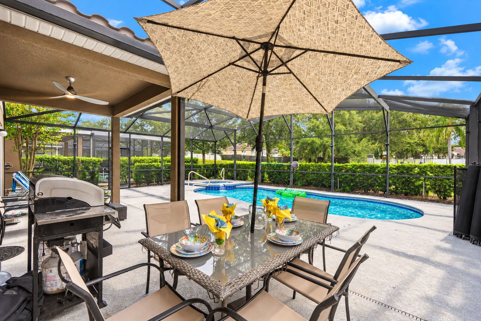 An outdoor patio with a glass table, chairs, a grill, and a pool in the background.
