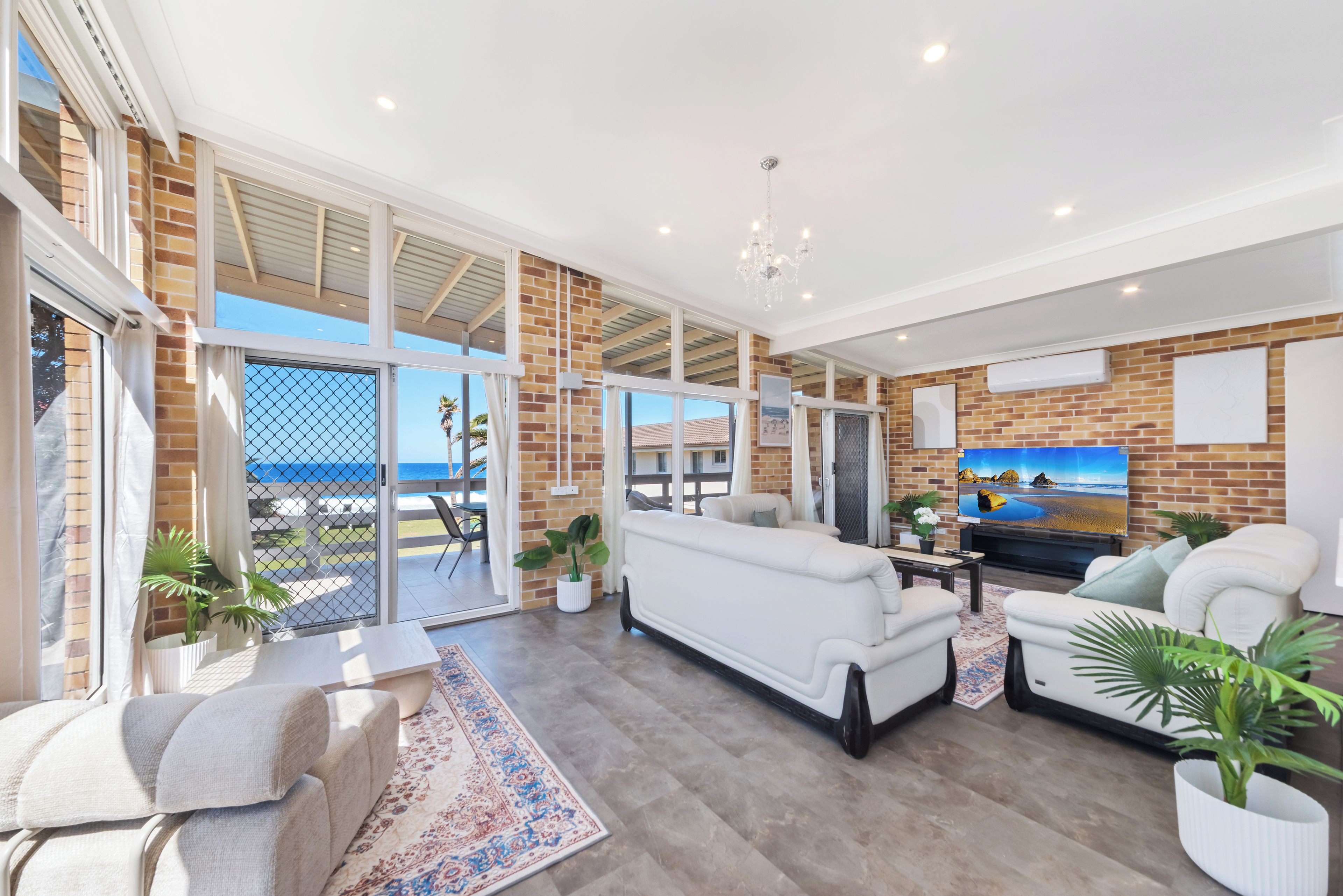 A spacious living area with a large sofa, a television, and a view of the beach through the glass doors.
