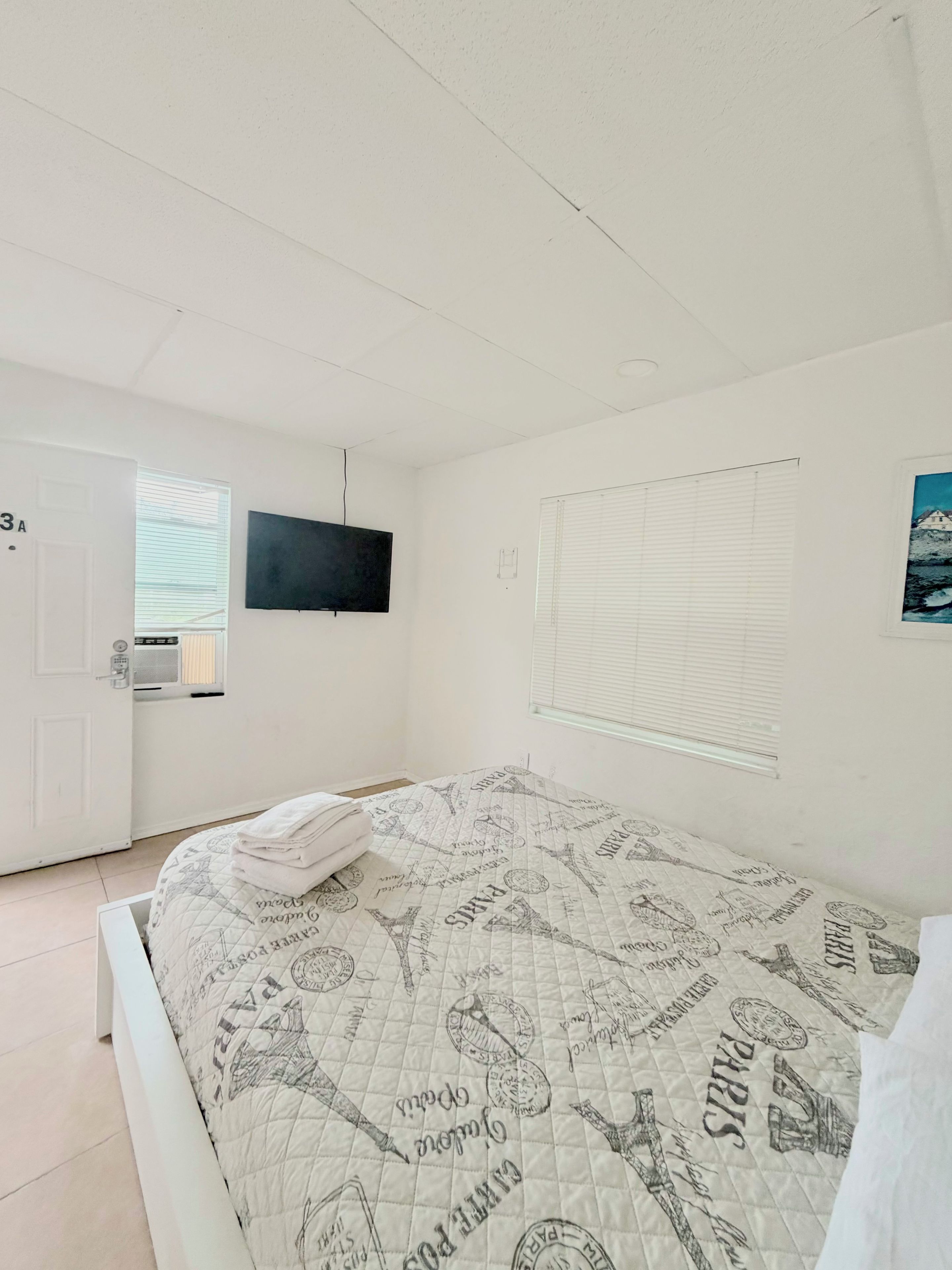 A room with a bed, a TV mounted on the wall, a door, and a window with blinds.