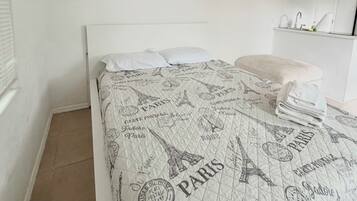 A bedroom with a bed featuring a Paris-themed quilt, a white headboard, and a small bedside table with a lamp.