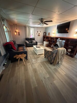 A living room with a ceiling fan, a black leather sofa, a patterned armchair, a white coffee table, and a television mounted on the wall.