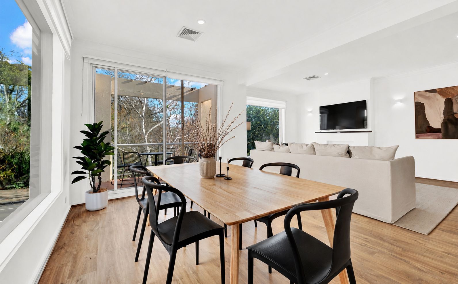 A modern living room with a wooden dining table, black chairs, a sofa, and a flat-screen TV.