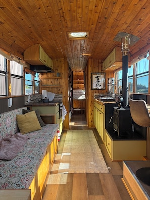 Interior acogedor de una cabaña con techo de madera, un sofá, una zona de cocina con estufa y una ventana que proporciona luz natural.
