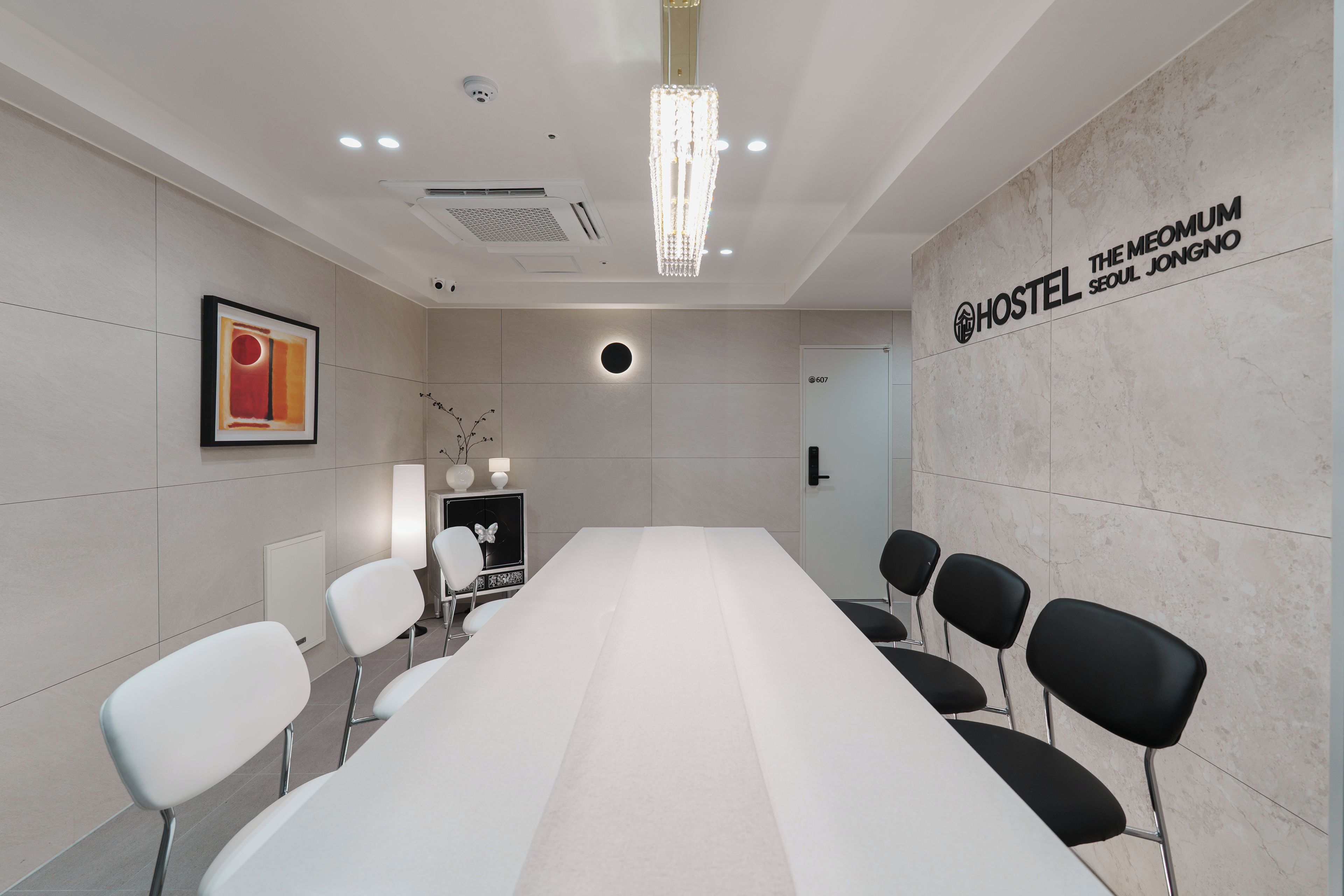 A conference room with a long table, chairs, a chandelier, a framed picture, and a wall sign for 'HOSTEL THE MEOMUM SEOUL JONGNO'.