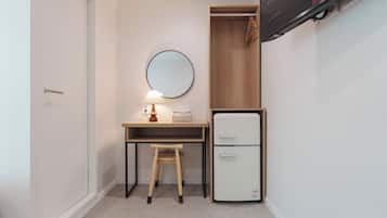 A compact room with a desk, a small stool, a mirror, and a white mini-fridge.