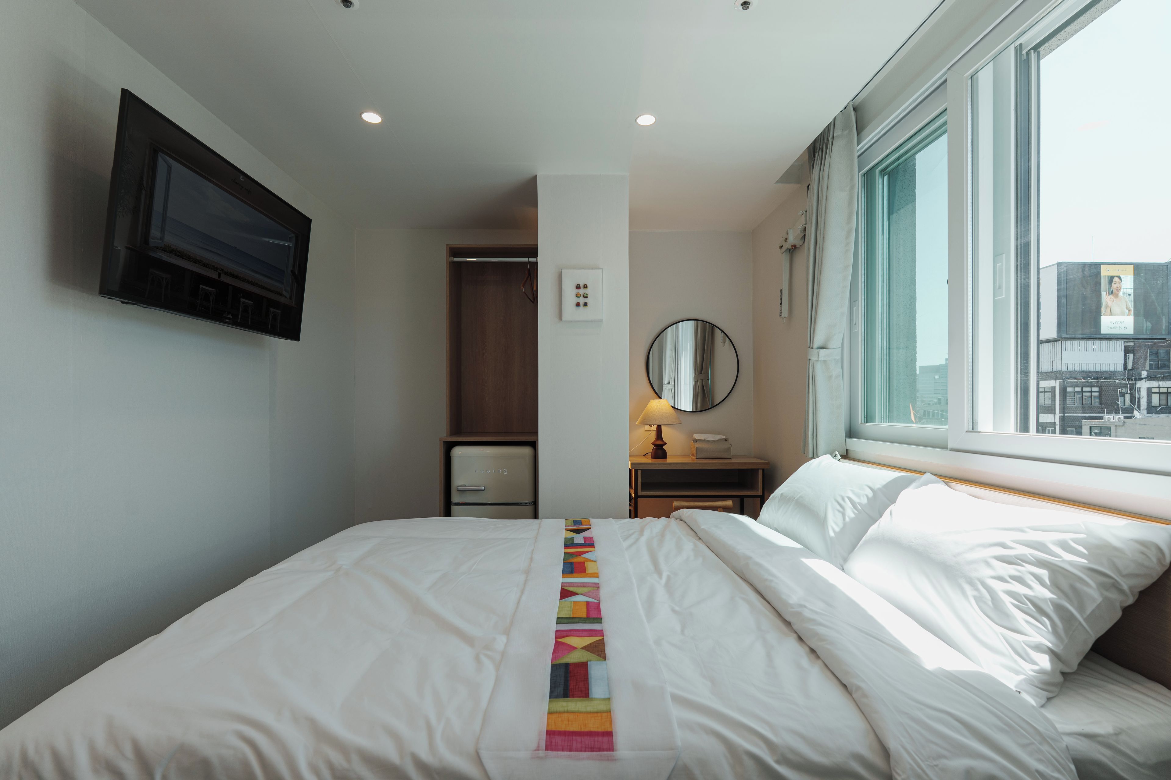 A hotel room with a bed, a bedside table with a lamp, a mirror, a wardrobe, and a TV mounted on the wall.
