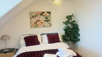A neatly made bed with a dark red blanket and white pillows, a wall-mounted painting, a potted plant, and a modern chandelier.
