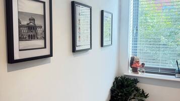 A hallway with framed pictures on the wall, a window with blinds, and a potted plant.