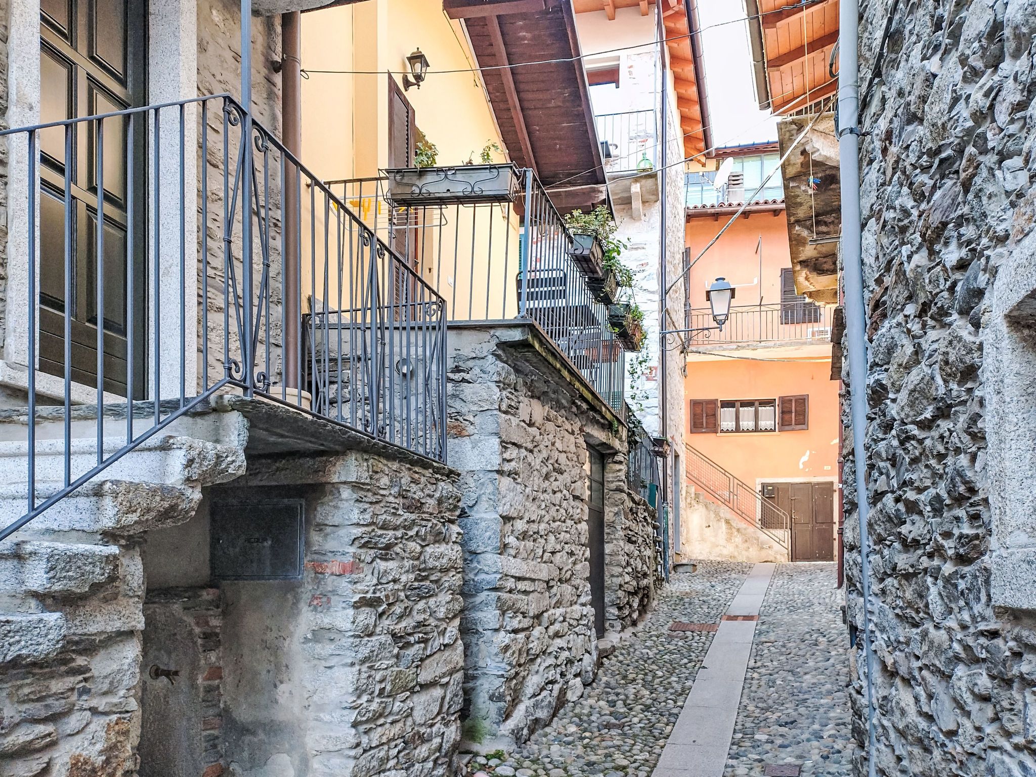 A narrow alleyway with stone walls, a metal railing, and a small balcony with potted plants.