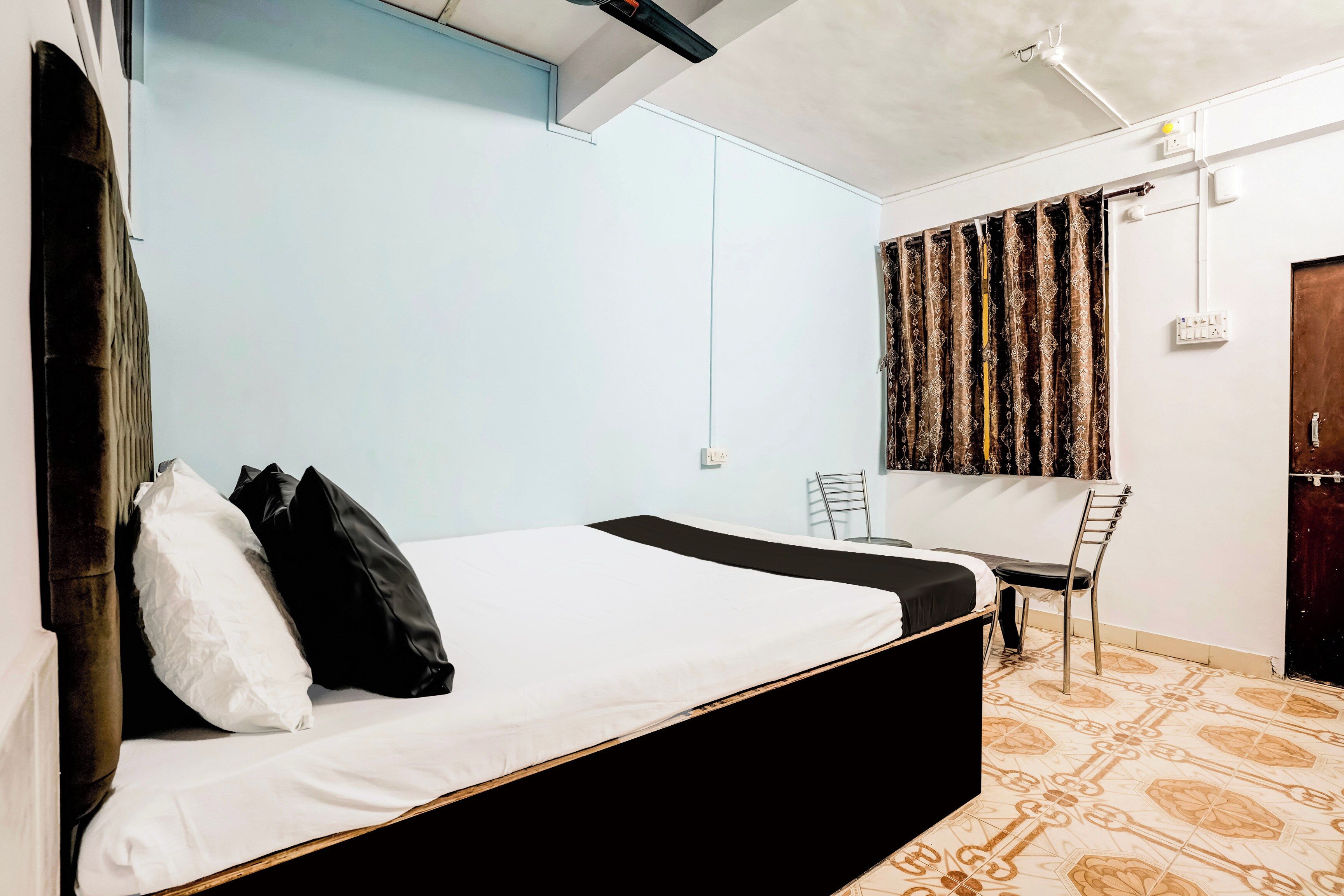 A single bed with a black frame, white bedding, and black pillows. There is a chair and a desk with a chair, both against the wall. The room has light blue walls and a patterned floor.