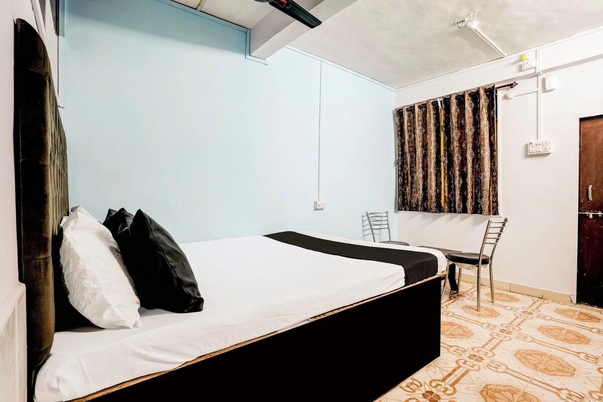 Una cama individual con estructura negra, ropa de cama blanca y almohadas negras. Hay una silla y un escritorio con silla, ambos adosados a la pared. La habitación tiene paredes azul claro y suelo con estampado.