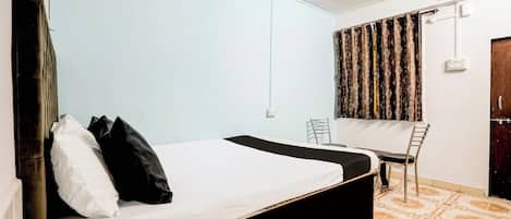 Una cama individual con estructura negra, ropa de cama blanca y almohadas negras. Hay una silla y un escritorio con silla, ambos adosados a la pared. La habitación tiene paredes azul claro y suelo con estampado.