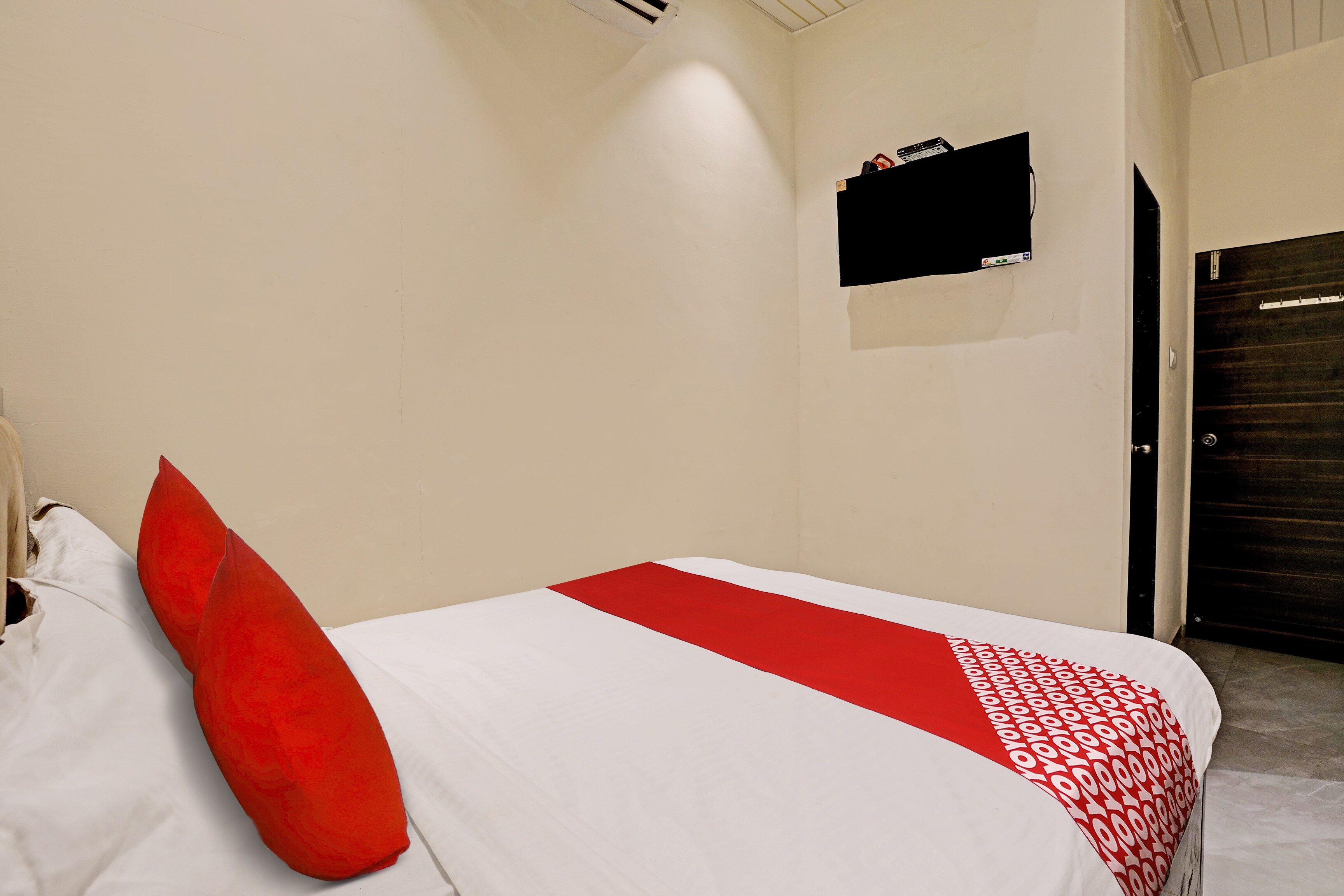 Habitación de hotel con una cama, almohada roja, colcha blanca y roja, una ventana pequeña con un objeto negro, y una puerta.
