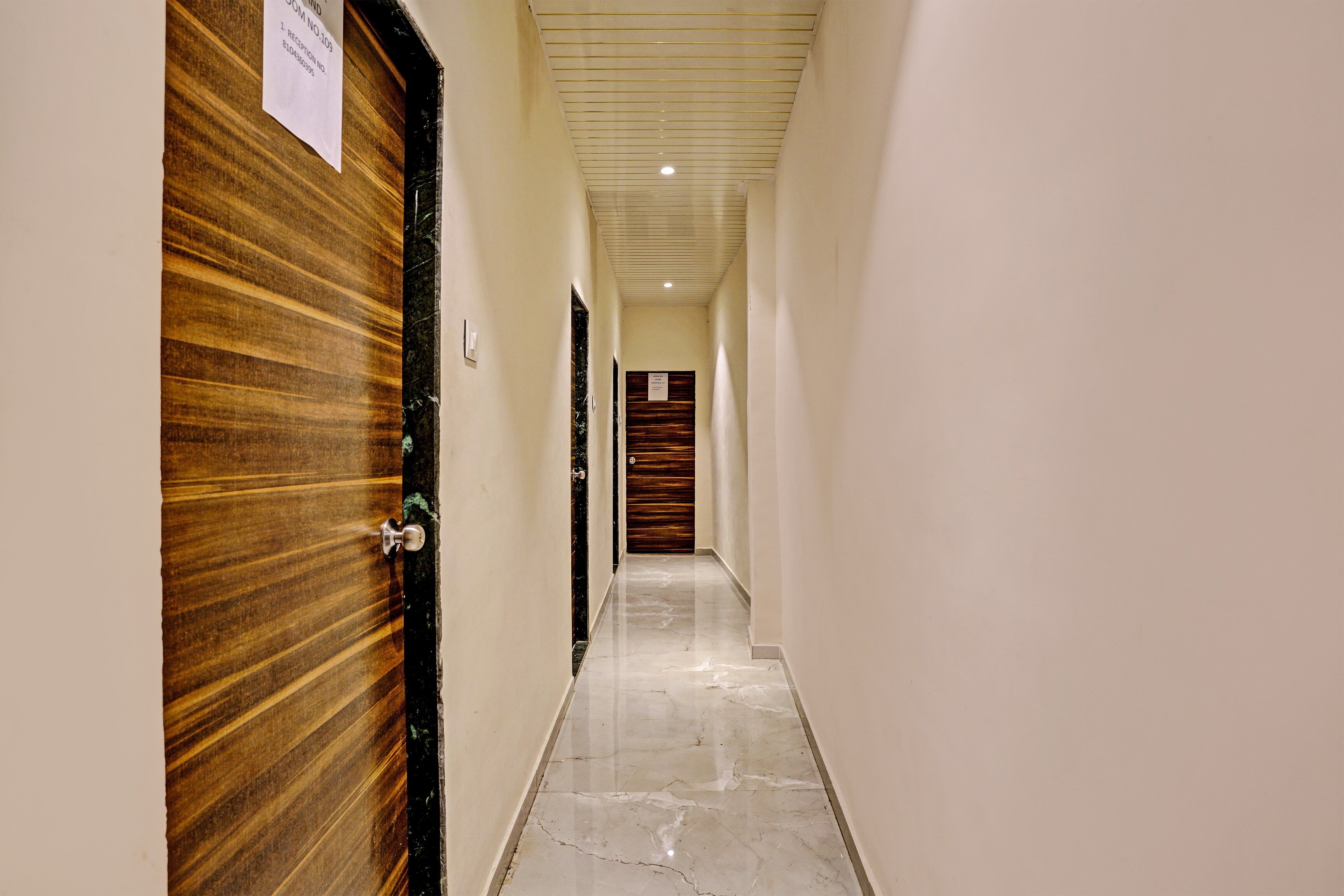 A corridor with wooden doors on the left, a marble floor, and a ceiling with recessed lighting.