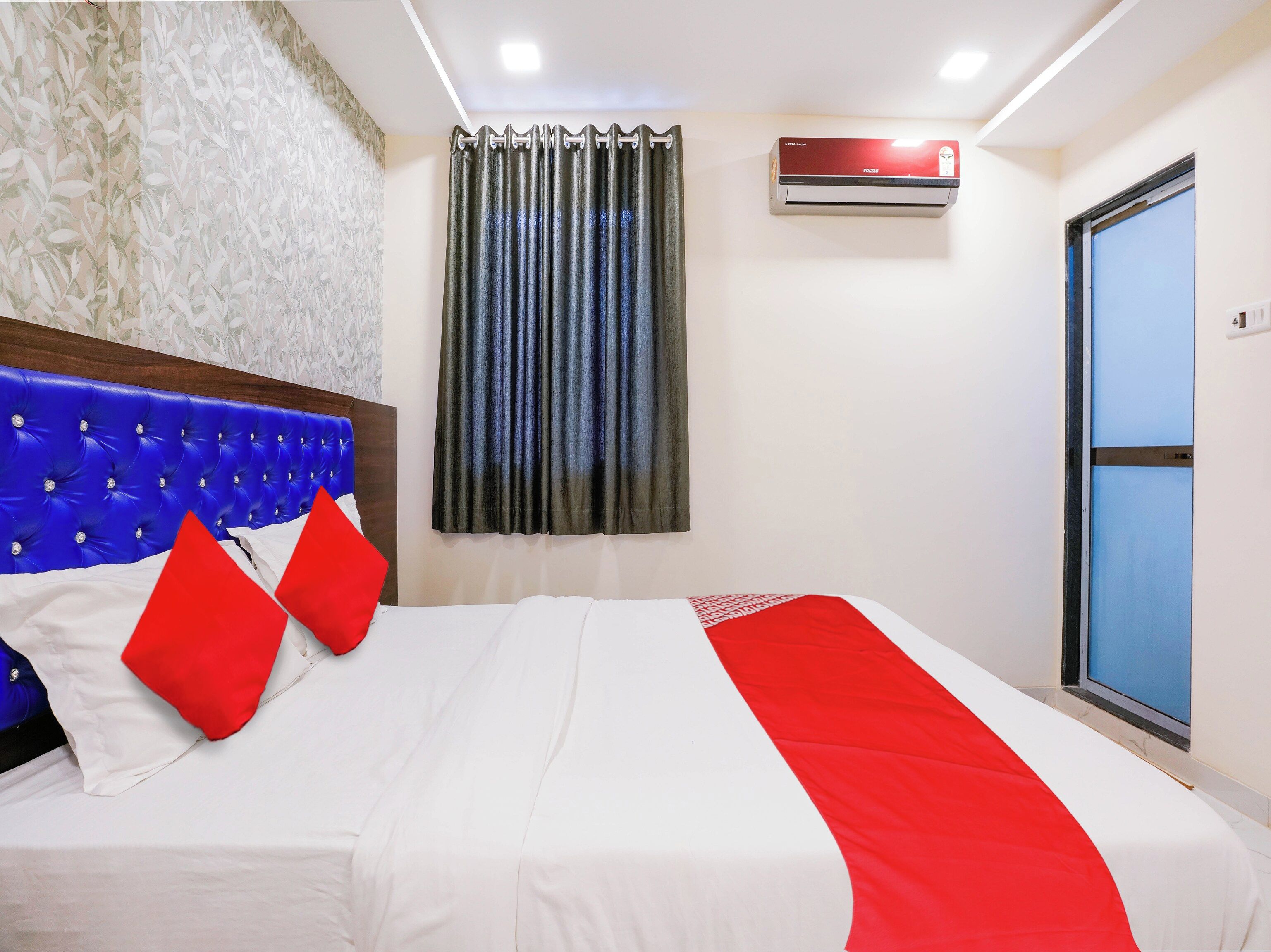 A hotel room with a bed, a headboard, a window with curtains, an air conditioning unit, and a door.