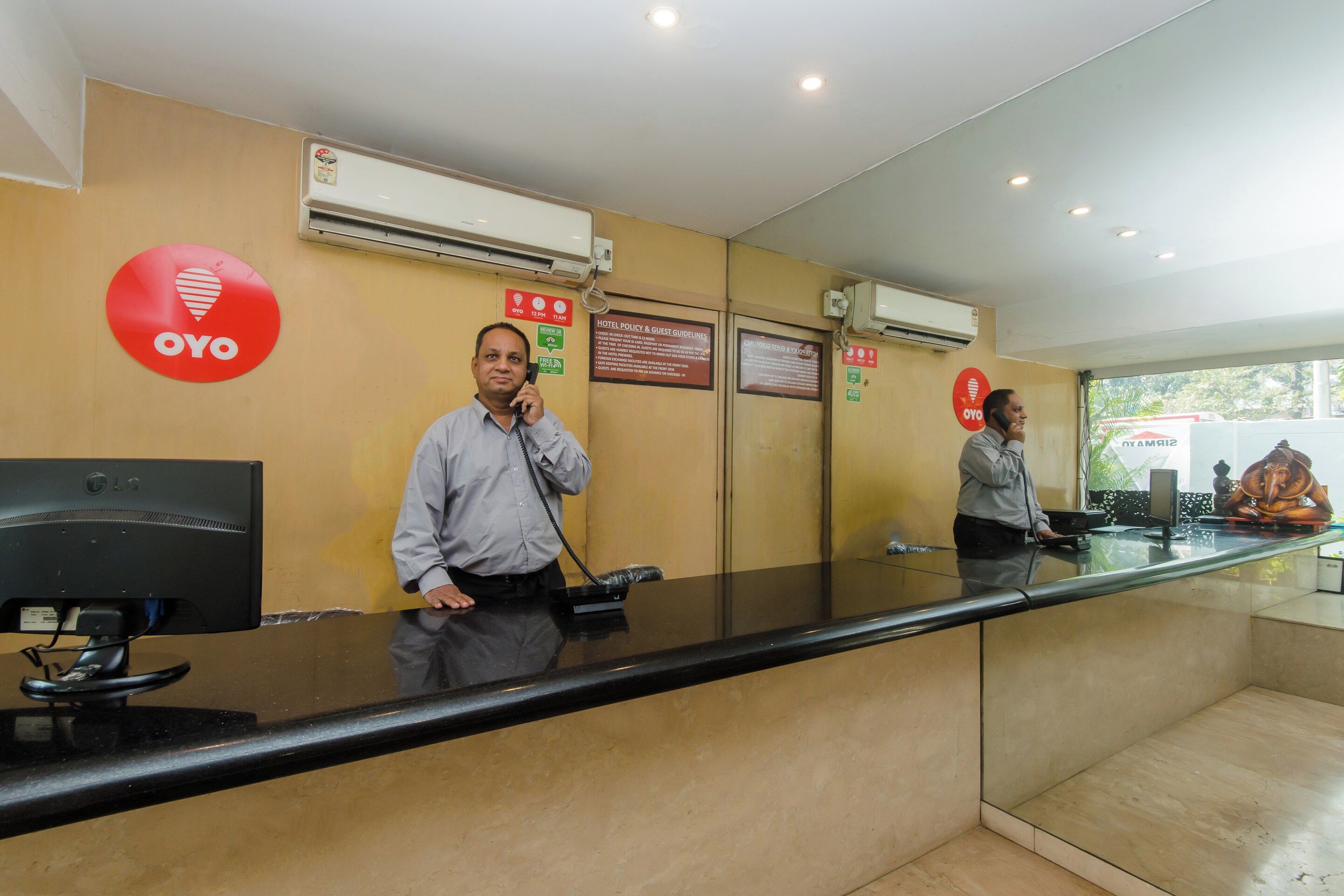 A hotel reception desk with a glass partition, a red OYO sign, and a sign indicating check-in times.